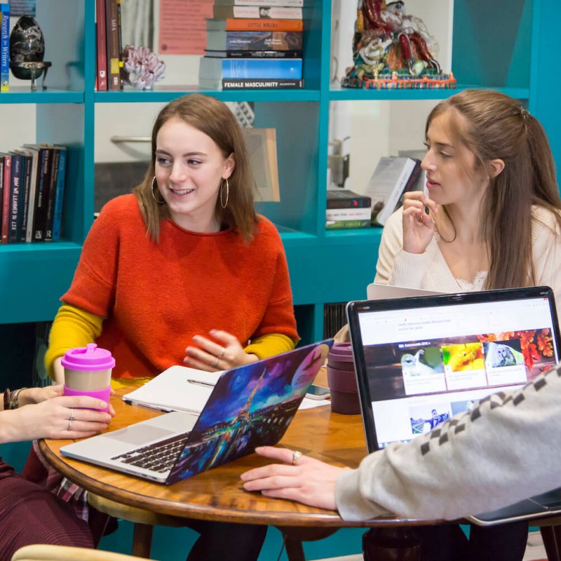 Four Falmouth University students talking at a table in front of bookshelves.