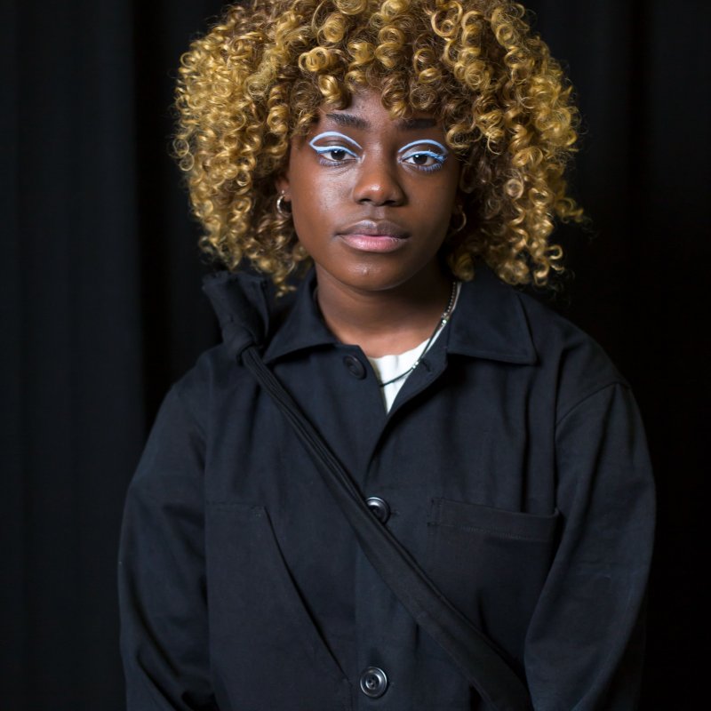 Student portrait - a black woman with curly golden hair poses in black clothing, with light blue eye liner