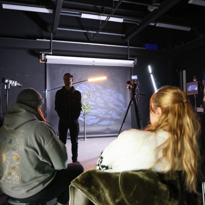 A dark room with a circle of students looking at a man presenting in the middle