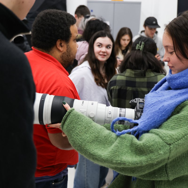 A person with shoulder-length dark hair holding a white camera lens, wearing a green knitted jumper and blue scarf