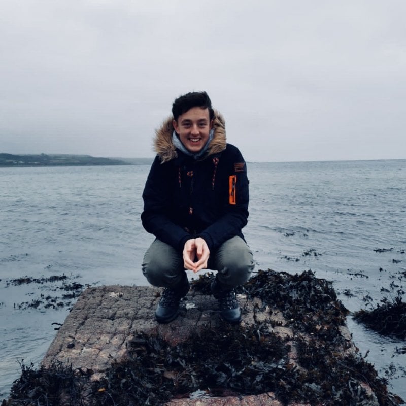 A male student crouching on rocks with sea in the background