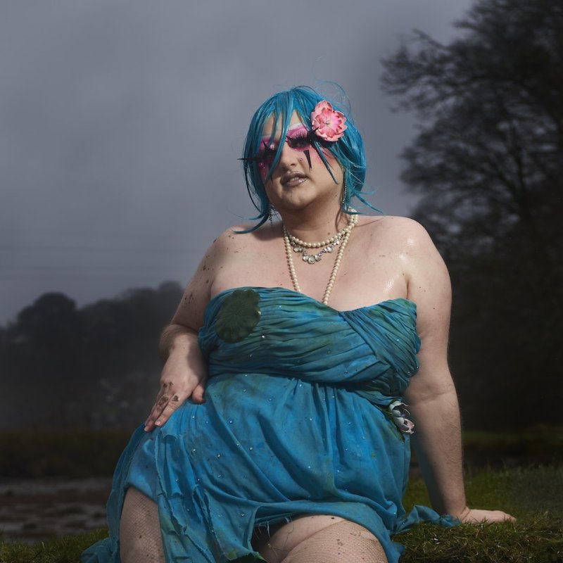 Illustration of a transgender person wearing a distressed blue dress, with blue hair, pearl necklaces and a pink flower in their hair