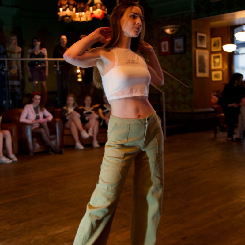 A woman poses on the catwalk wearing shiny black boots, olive trousers and a cropped white top. 