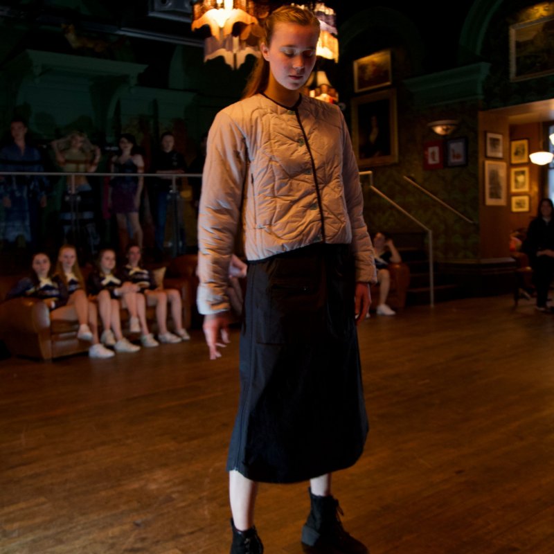 A student poses on the catwalk. She wears suede black boots, a shin length black skirt and an off-beige jacket