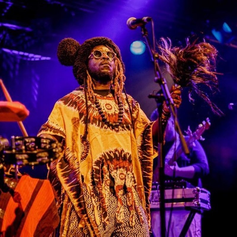A man with dreadlocks performing on a stage with a drum kit behind