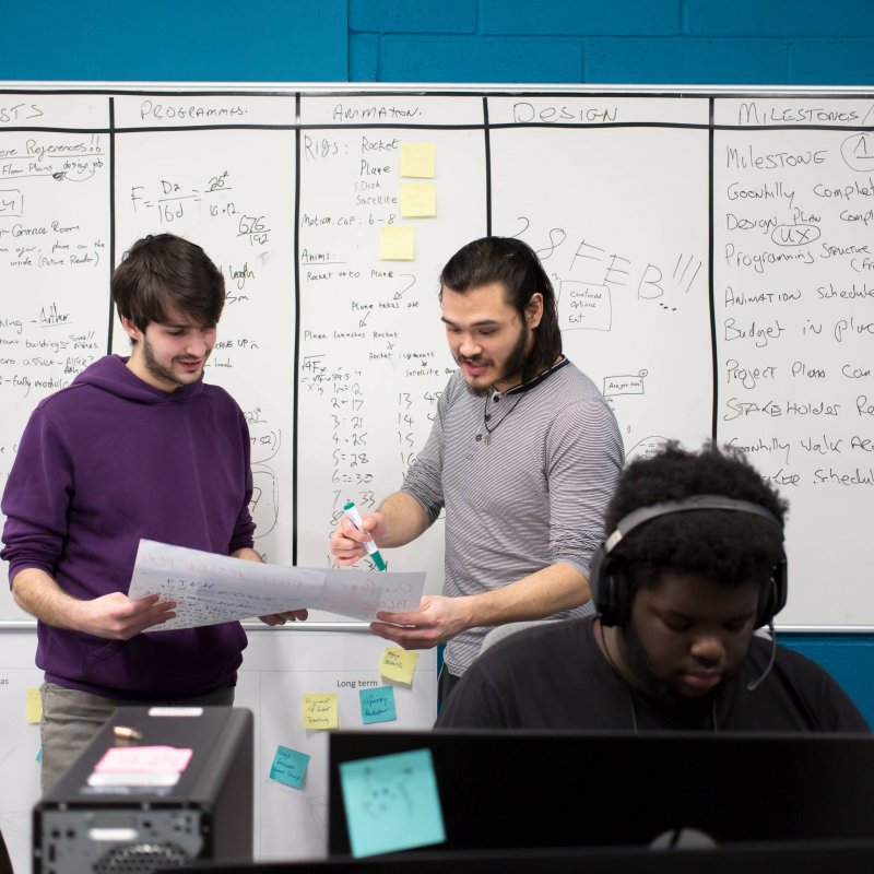 Two students stand in front of a whiteboard scrutinizing a piece of paper. A student sits in the foreground working on a laptop. 