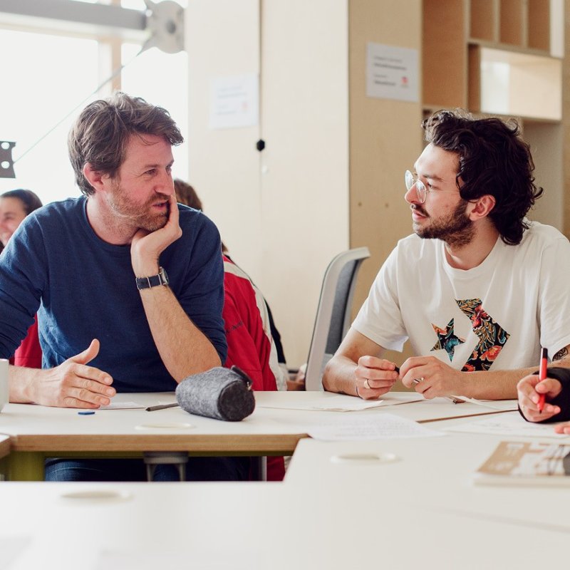 Two men talking to each other while sat at a table.