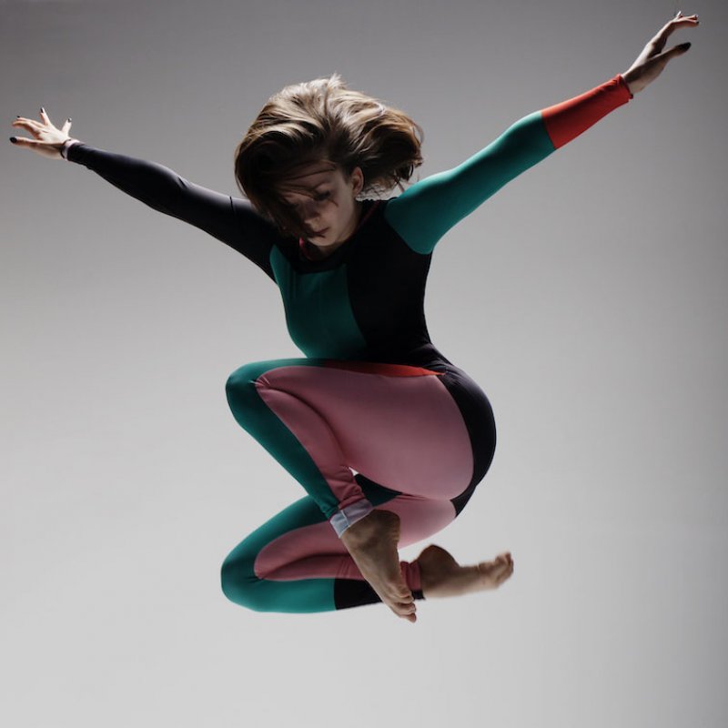 Dancer jumping high with arms spread and legs tucked under in grey backdrop studio.