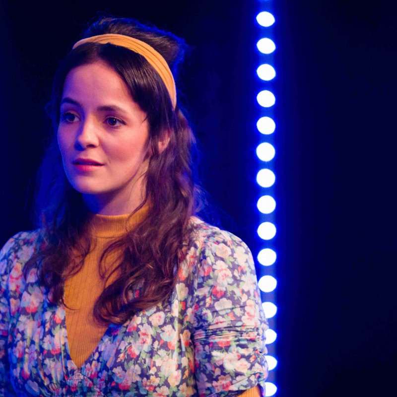 A woman dressed in a flowery top stands in front of blue lights