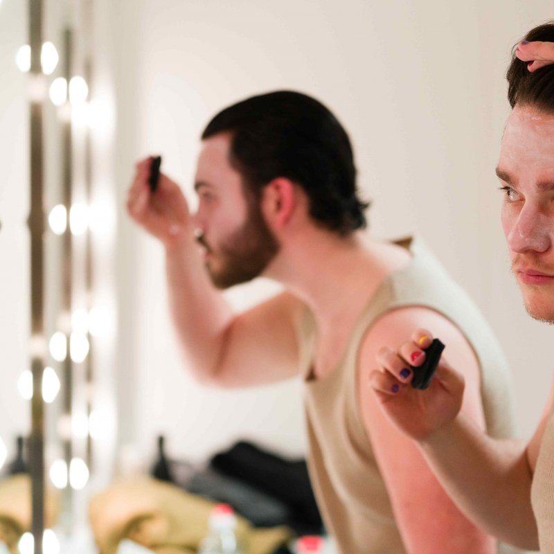 Two men look into mirrors, doing their makeup before a performance