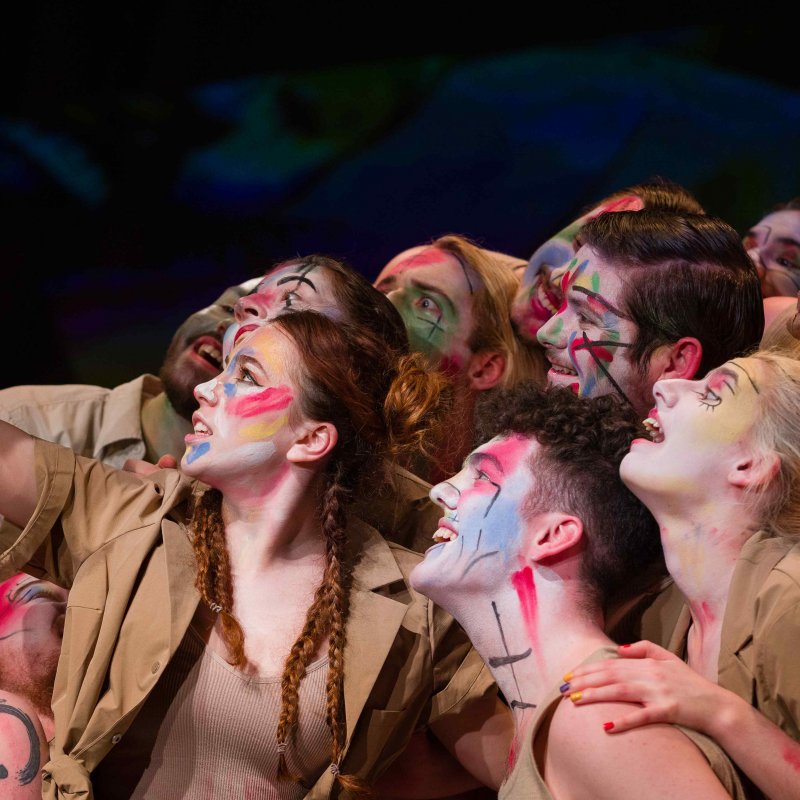 A group of actors with painted faces look towards a hand-held tablet 
