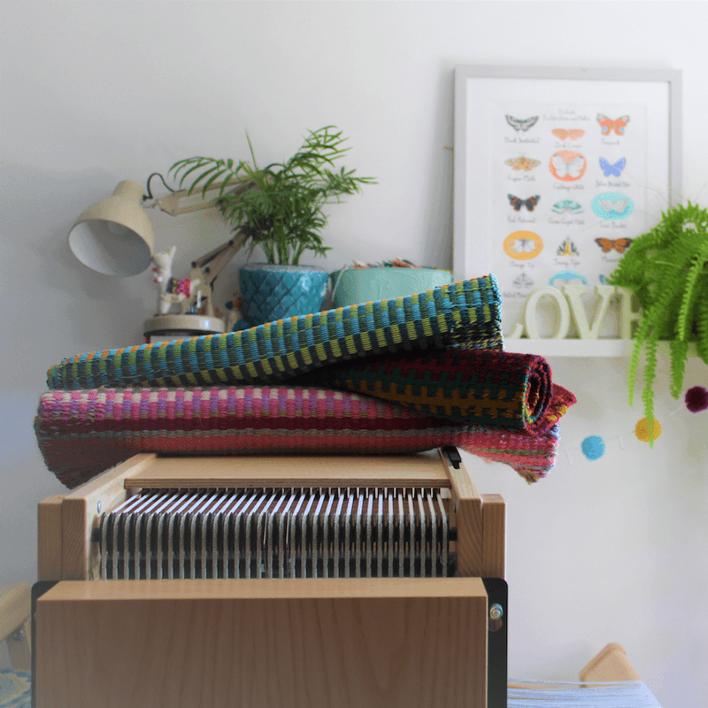 Rolled up woven fabric on top os a loom with a plant and lamp in the background