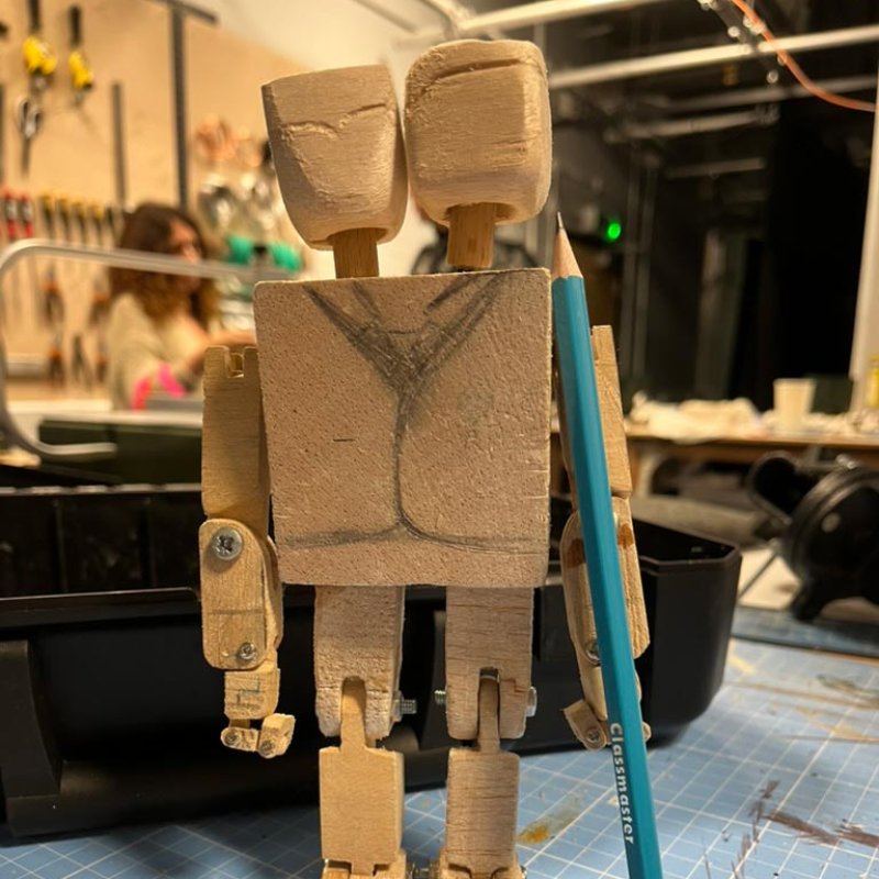 An unpainted wooden puppet with two heads next to pencil