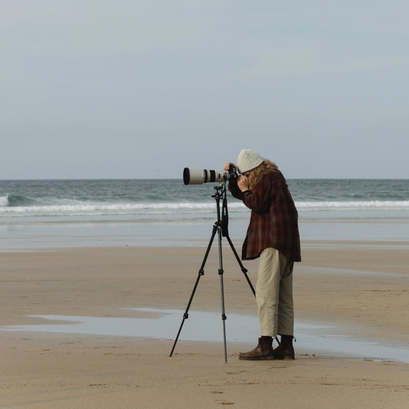 A woman standing on the beach using a large camera on a tripod, 