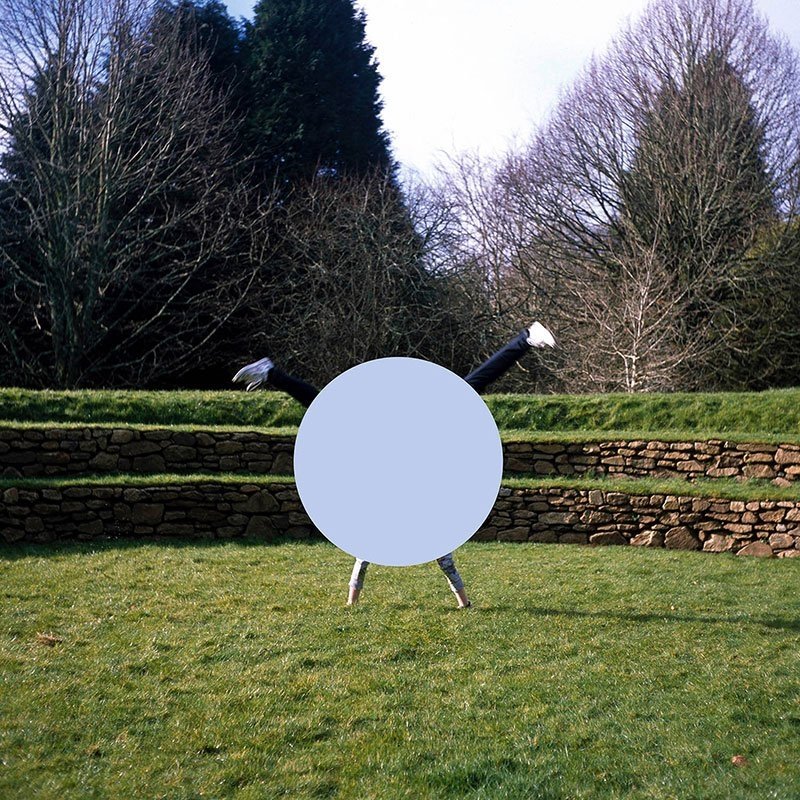 Photo of someone doing a handstand on a green lawn, with trees and grey sky in the background. A light blue circle is covering the person so that only arms and legs are showing.