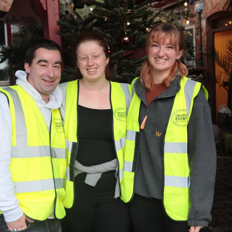 Three people posing together wearing jumpers and yellow hi vis jackets with Christmas tree and lights in the background