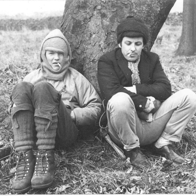 Two men wearing winter clothes, smoking and sitting by a tree