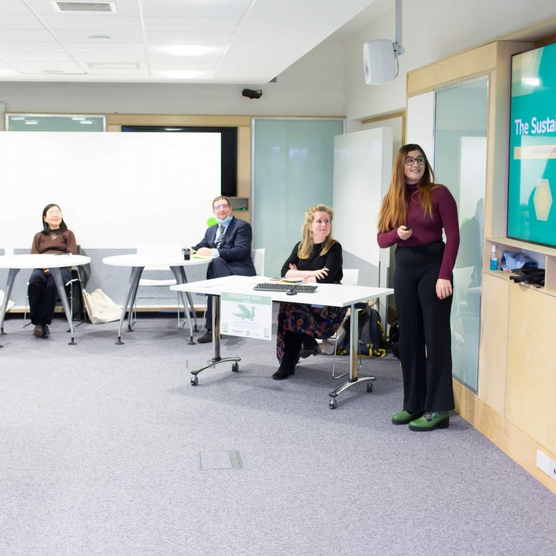 A student stands in front of a television screen and presents their business pitch to a panel of experts