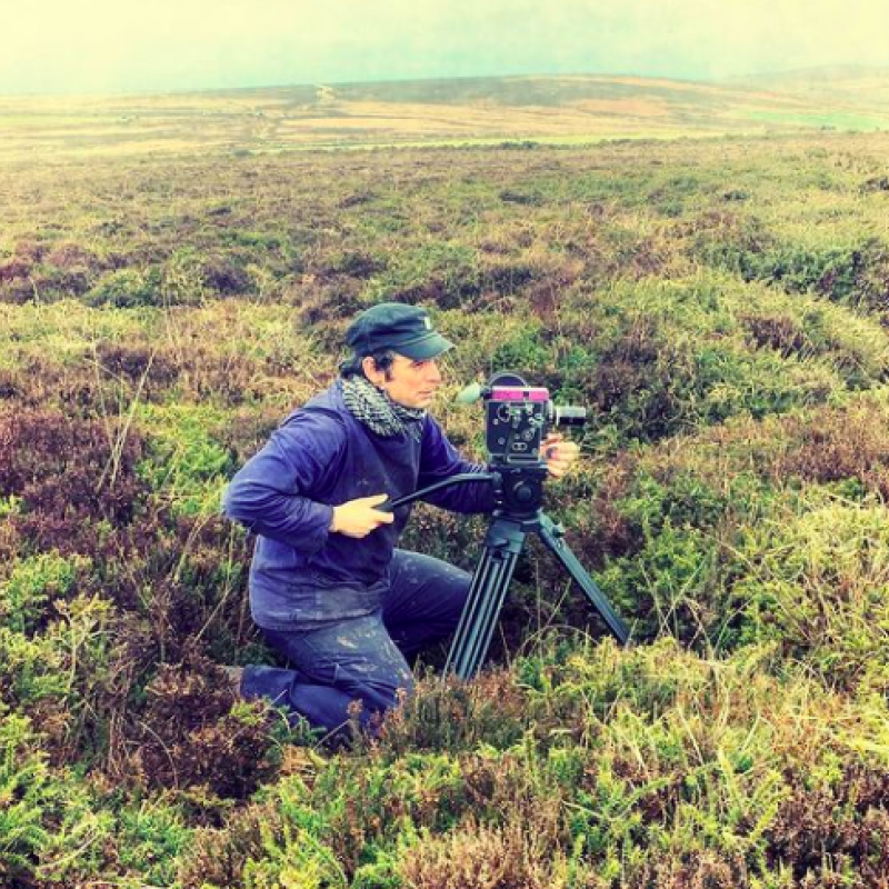 A filmmaker crouches behind his camera amongst shrubbery 