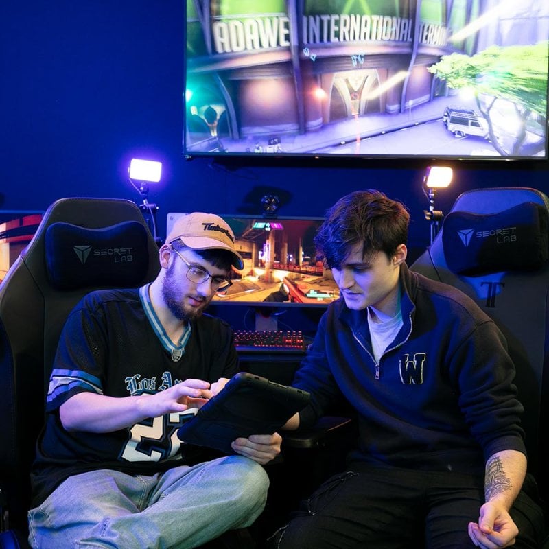 Two male students looking at an iPad with large gaming screens behind