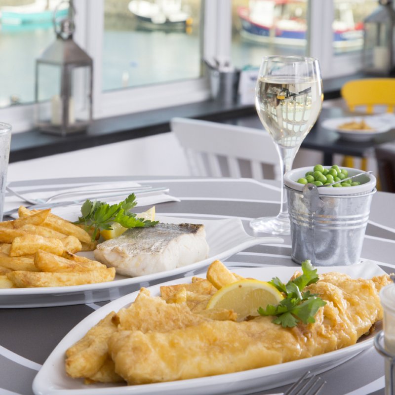 Two plates of fish and chips with a bucket of peas and drinks on a table