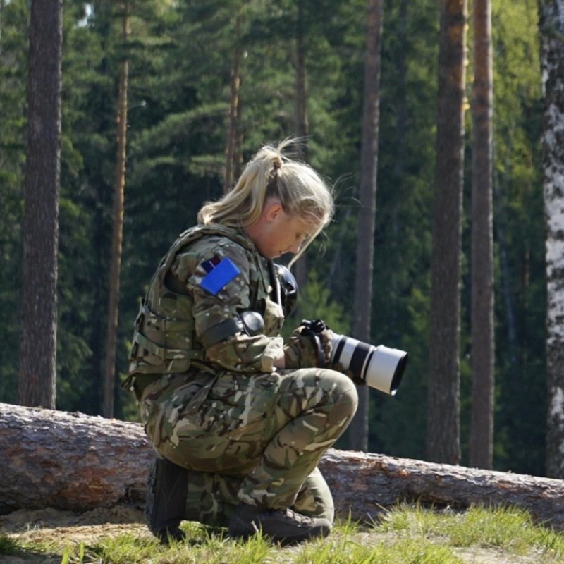 A woman dressed in camouflage uniform, kneeling and holding a camera