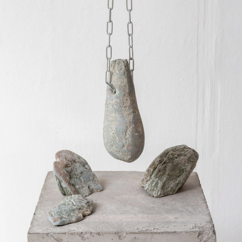 Three stones are arranged on a stone plinth. A fourth stone hangs above the ensemble from a silver chain