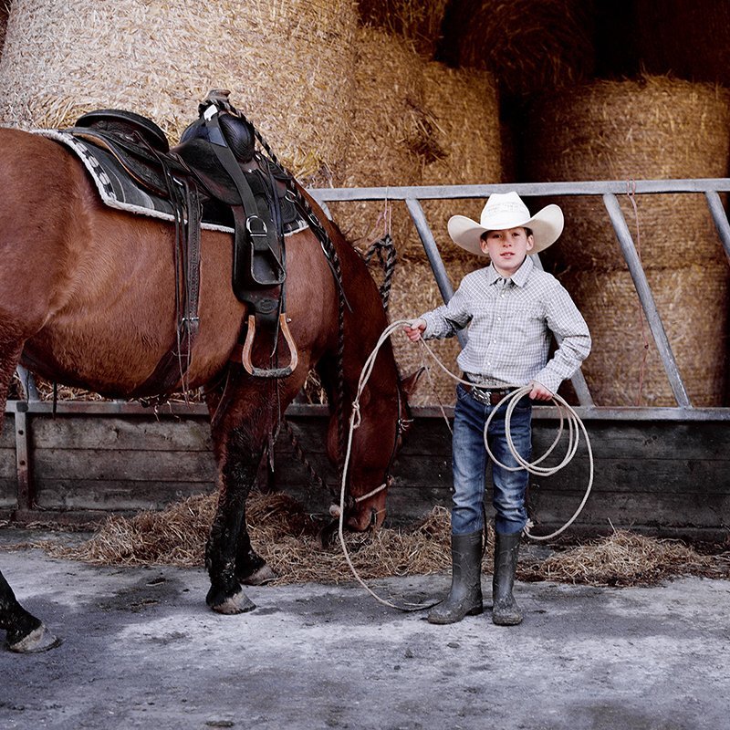 Photo of a young boy - wearing a shirt, jeans, wellington boots and cowboy hat - holding a lassoo in front of a horse with hay bales in the background.