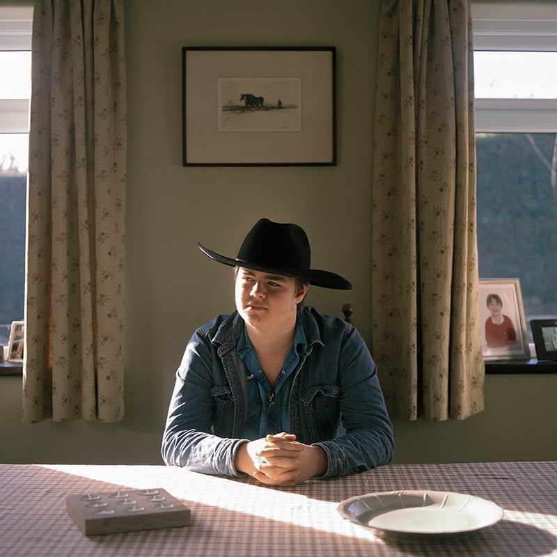 Photo of a cowboy, wearing a denim shirt and jacket and a brown cowboy hat, sat at a table with a red and white table.