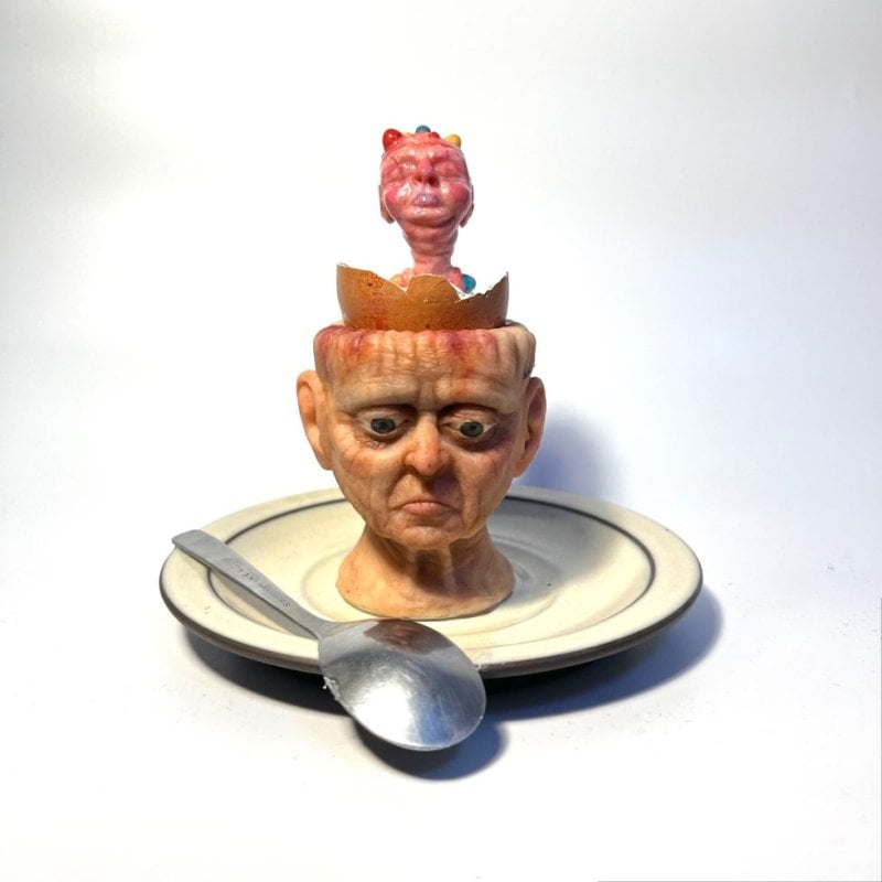 Photo of a miniature sculpture - an egg cup in the shape of a human head sat on a saucer. Inside the egg cup, a monstrous thing breaks out of its shell