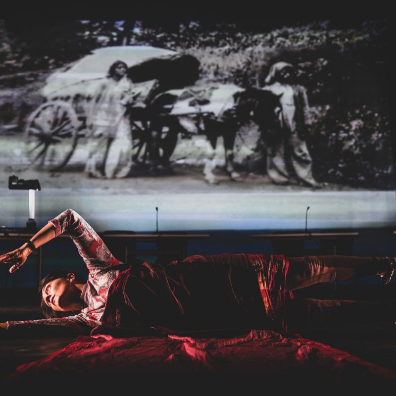 A woman lies on stage, covered in red velvet with a screen projection behind her
