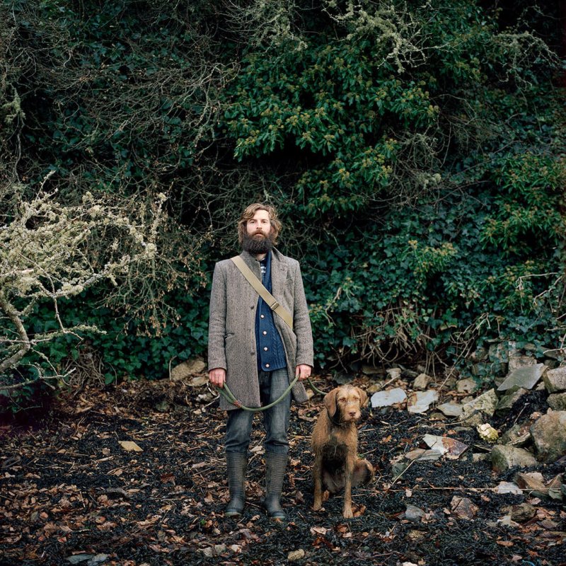 A man with a beard holding a dog with trees behind