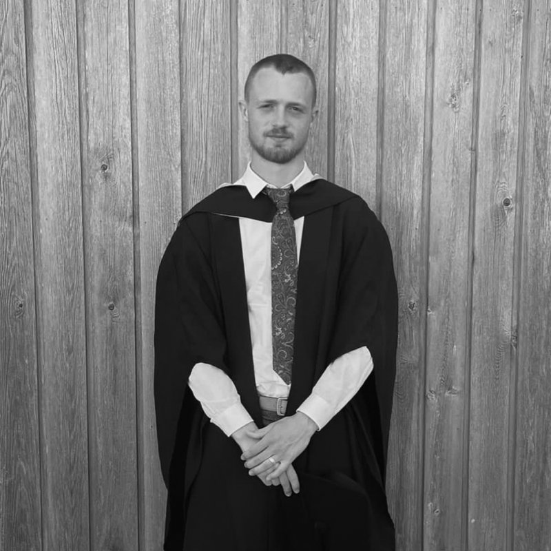 Student wearing graduation robe standing against wooden clad wall. 