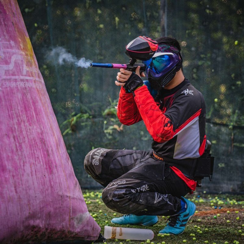 A man crouches wearing paintball gear, shooting a paintball gun 
