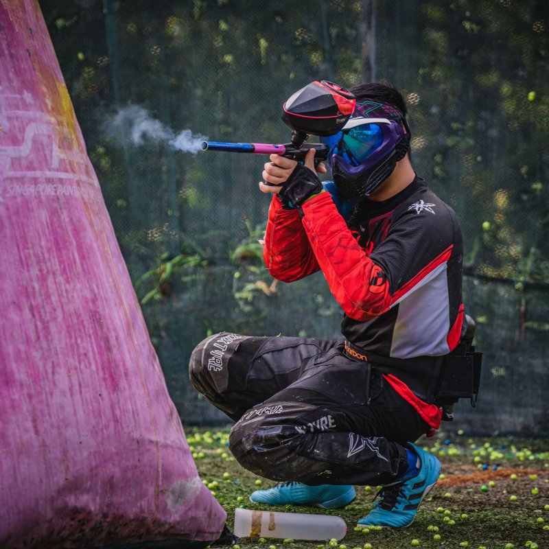 A man crouches wearing paintball gear, shooting a paintball gun