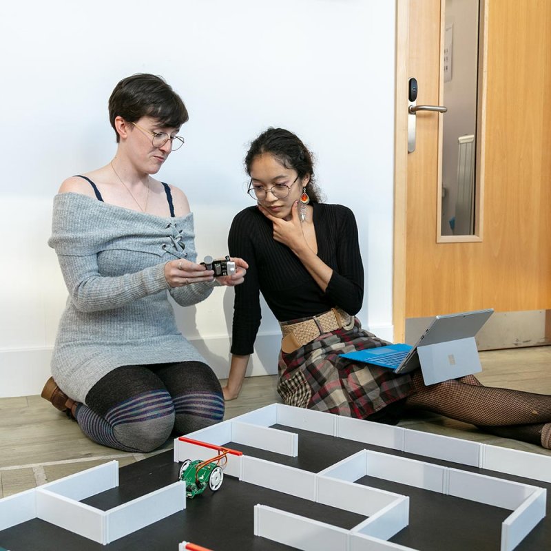 Two female Robotics students sitting on the floor examining a small robotic device