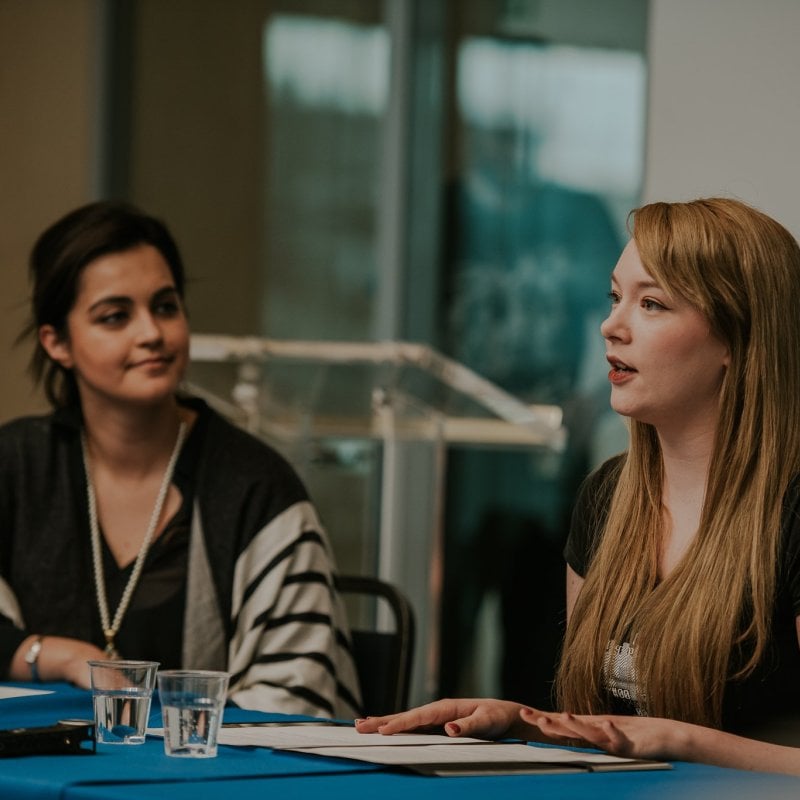 Two women sat next to each other while one gives a speech