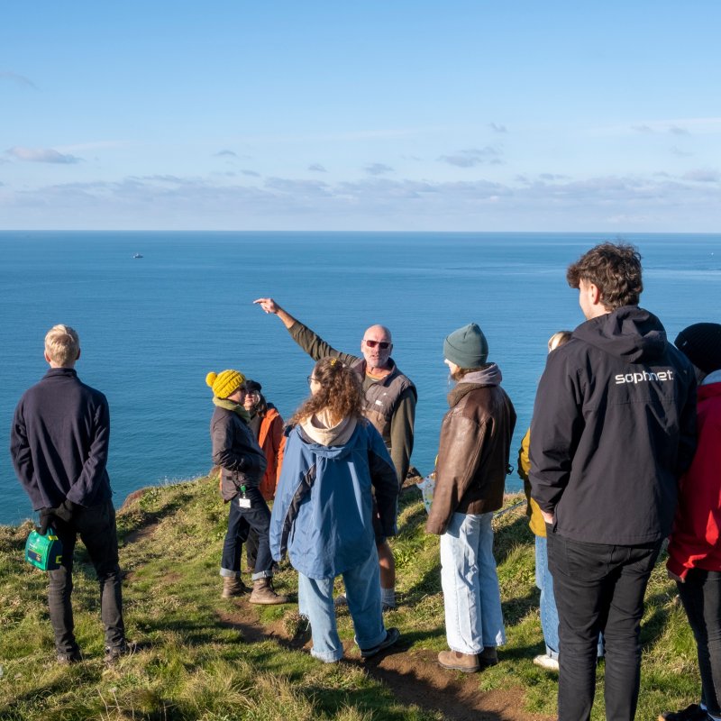 Artist Tony Plant stands on cliff top overlooking the sea with Falmouth University students during sand art project introduction
