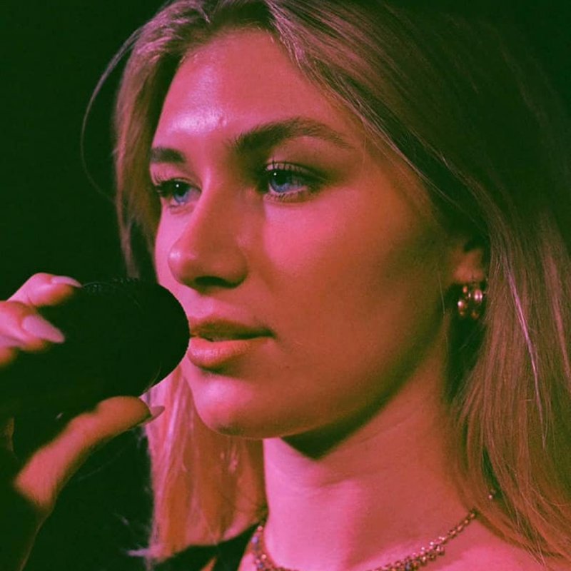 A close up of a woman singing into a microphone. She wears a gold necklace and is bathed in pink light