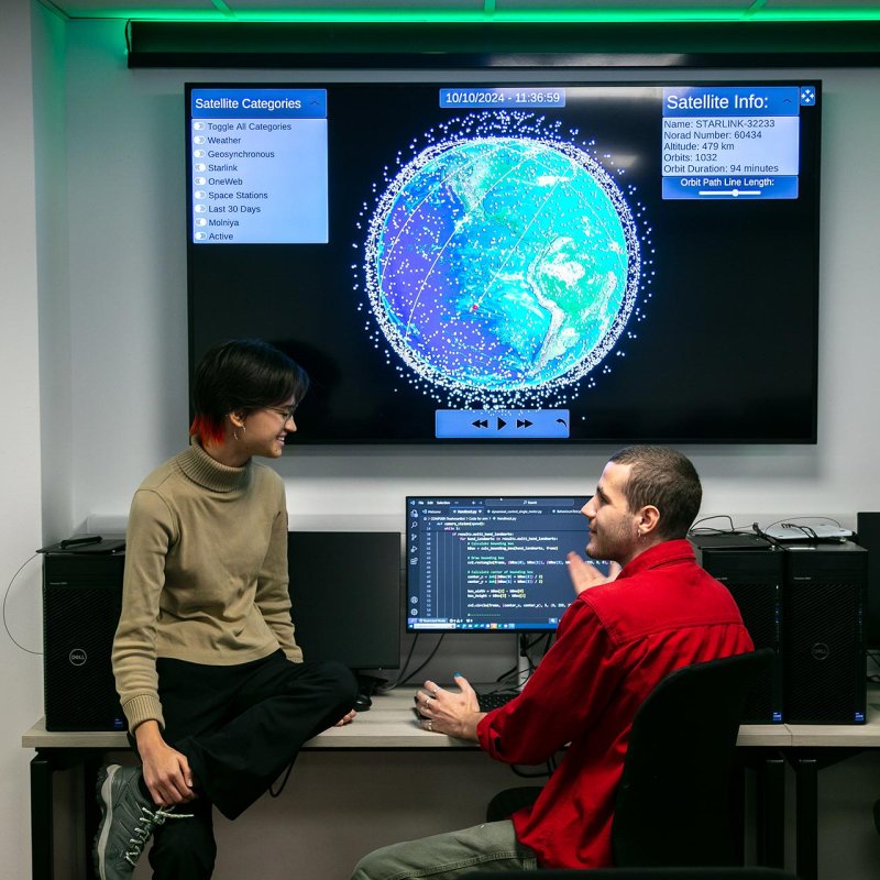 Two software engineering students in discussing with large screen of the world on the wall