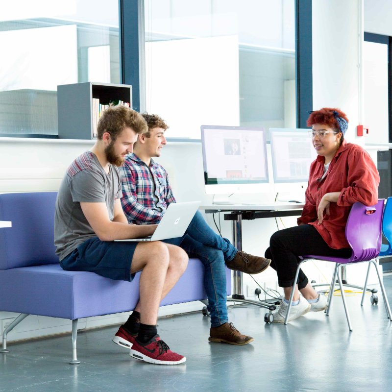 Two students sitting on a purple sofa next to another student at a computer desk, Falmouth University
