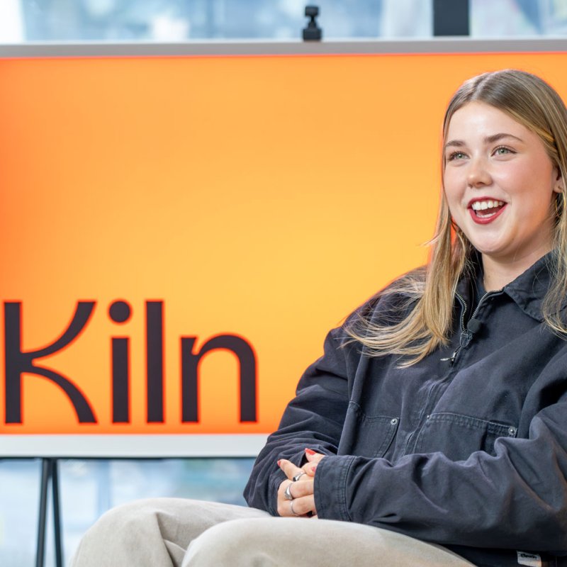 A Falmouth University graduate seated with orange sign saying 'Kiln' behind