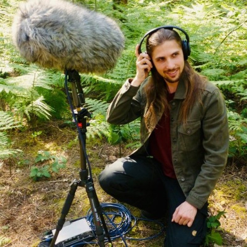 Photo of researcher Ton Hull crouched on the ground in woodlands wearing headphones in front of a microphone boom mic