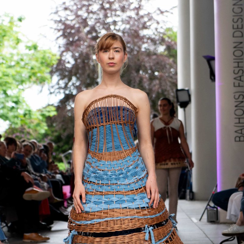 A woman wearing a wicker dress and blue ribbon on the catwalk