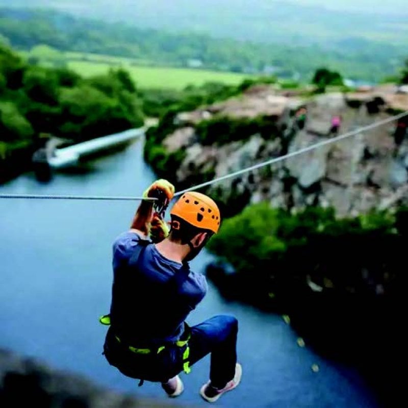 A young man on a zip wire handing above a large lake with trees in the background