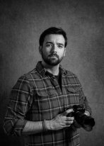 A black and white photograph of Alex Mighall holding a camera