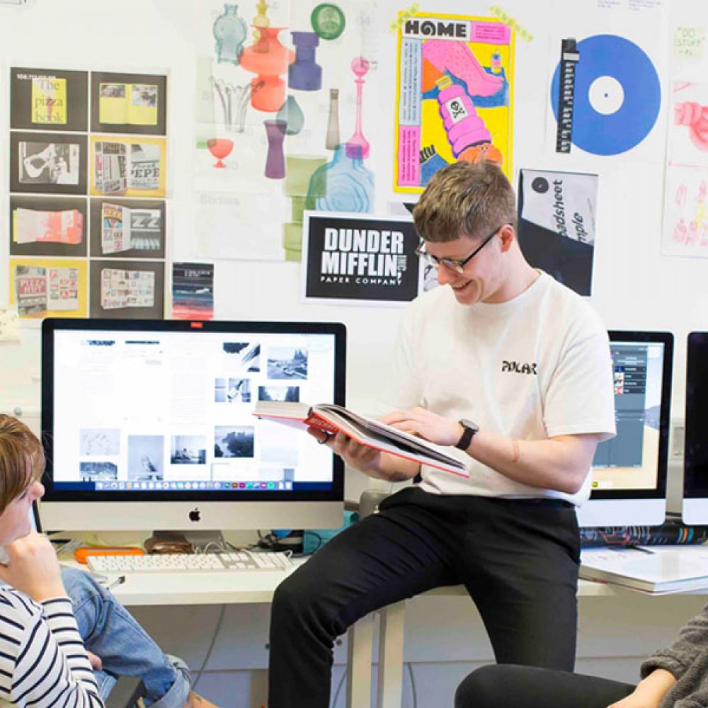 Three Falmouth University students sat round an iMac with graphic design posters on the wall