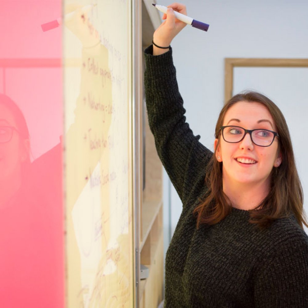 A woman wearing a black jumper, writing on a whiteboard with a pink panel