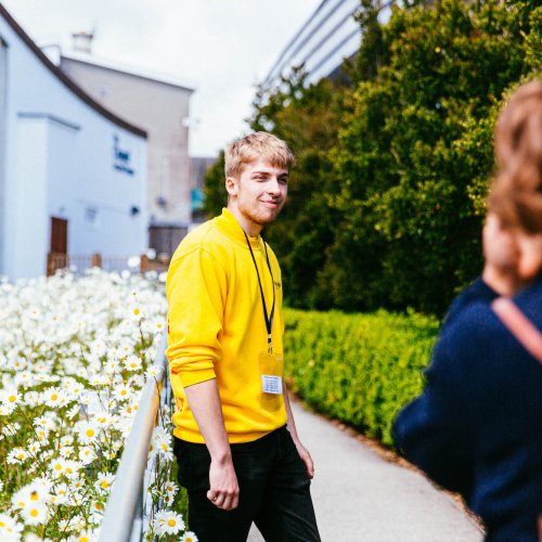 Male student wearing a yellow sweatshirt outside with daisies