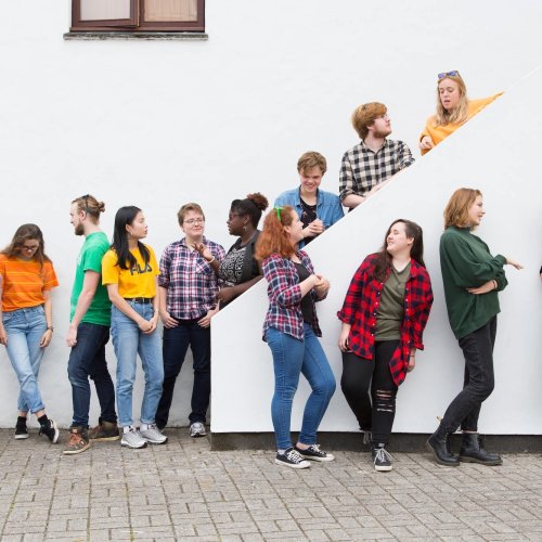 13 Falmouth University students in different coloured tops stood against a white wall and on a white staircase.